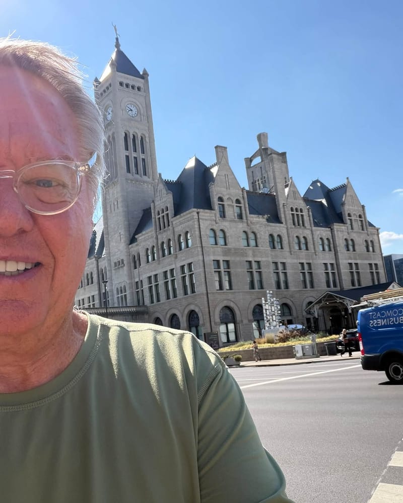 Man in olive green shirt taking selfie in front of historic Union Station hotel building with clock tower under clear blue sky