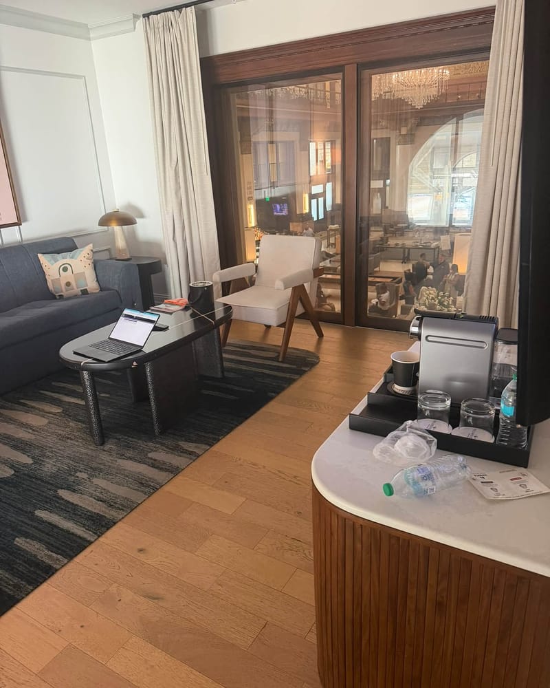 Hotel suite living area with gray sofa, laptop on coffee table, cream accent chair, wood floors, and large windows overlooking grand lobby with chandeliers