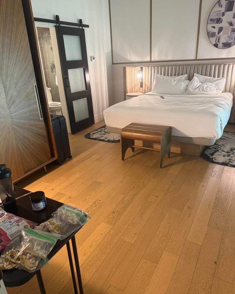 Spacious hotel bedroom with king bed in white linens, sliding barn door to bathroom, wood floors, modern decor, and luggage by closet